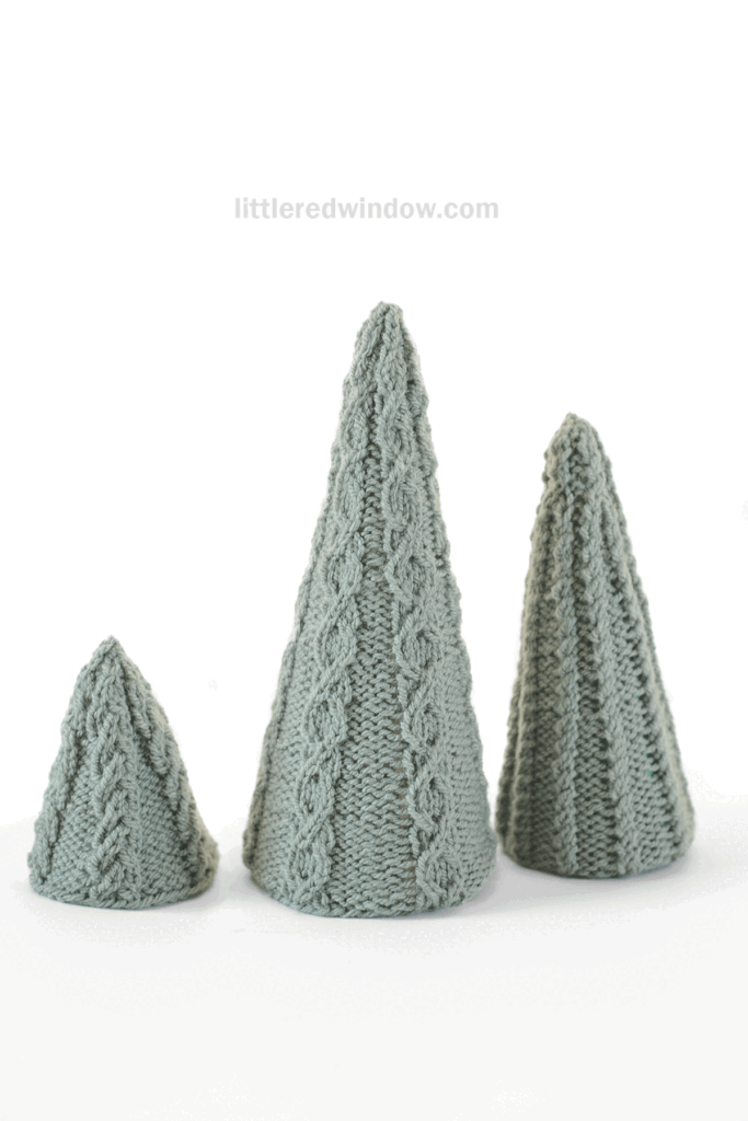 Three knitted cone-shaped cabled Christmas Trees in light gray-green yarn, each adorned with a cable knit pattern, are displayed on a white background.