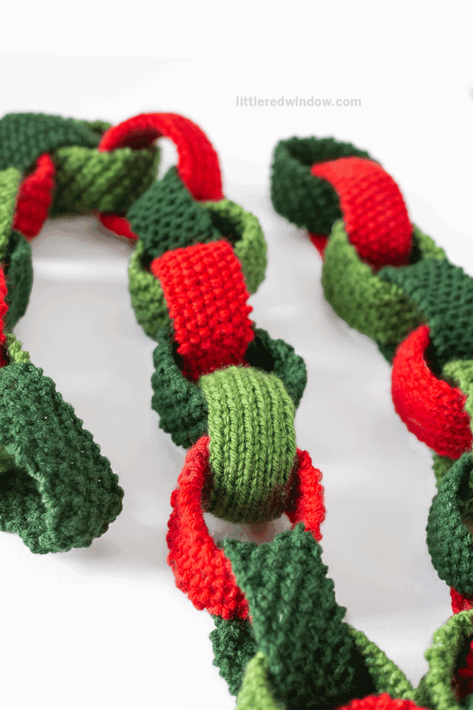 Close-up of a handmade garland, featuring red and green knitted chain links interlocked together, crafted using a Christmas Paper Chain knitting pattern, photographed against a white background.