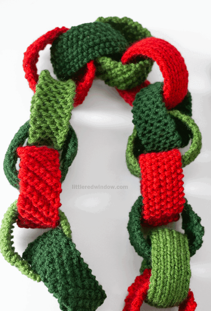 A close-up of a festive garland made from knitted links in red, dark green, and light green yarn, connected in a chain pattern inspired by a Christmas Paper Chain knitting pattern against a white background.