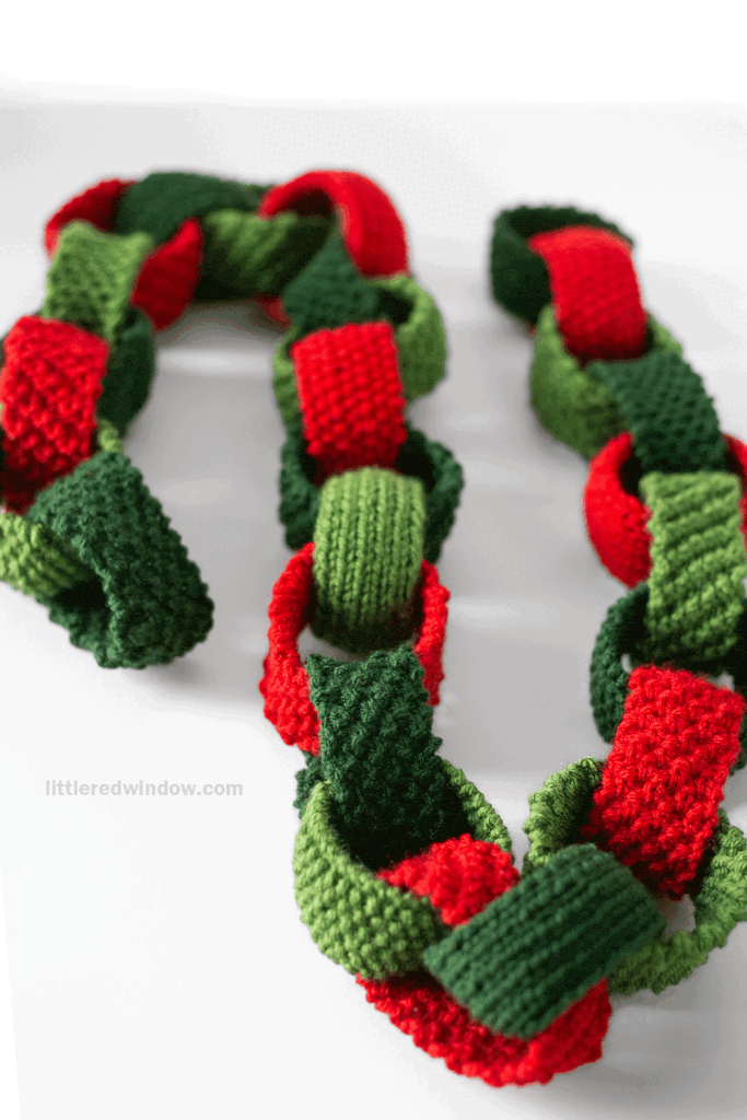 A knit garland inspired by the Christmas Paper Chain knitting pattern, featuring interlocking rings in red, dark green, and light green yarn, arranged in a loose, overlapping pattern on a white surface.