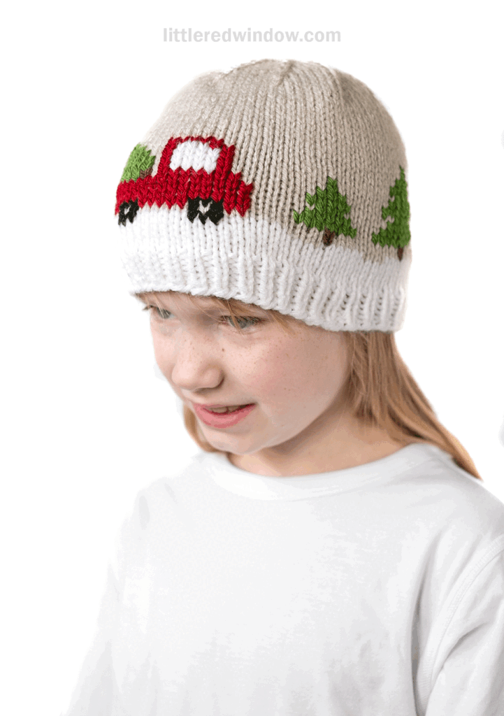 A child wearing a beige and white knit hat adorned with a christmas tree truck, smiling and looking slightly down while dressed in a plain white shirt.