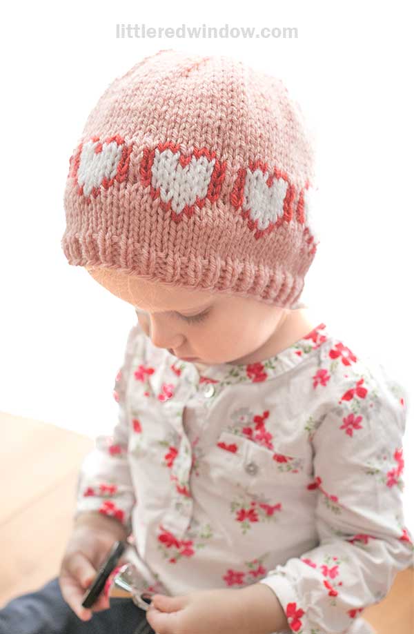 A toddler cozily sits indoors wearing a heart hat, its pink knit adorned with white patterns. The child focuses intently on something in their hands, dressed in a charming white floral patterned shirt.