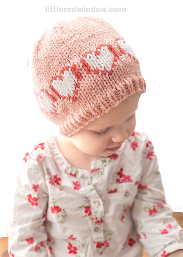 A child wearing a heart hat in pink knit with white patterns looks downward. The floral-patterned, button-up shirt adds charm against a soft white background.