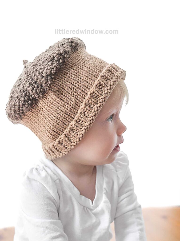A baby wearing an Acorn Hat sits and looks to the side. The hat is brown with a darker brown top, mimicking an acorn cap, and the baby is dressed in a white shirt. The background is plain white.