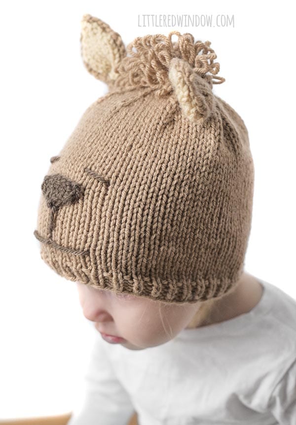 little girl looking down and to the left and wearing tan alpaca hat with brown nose and curly hair on top