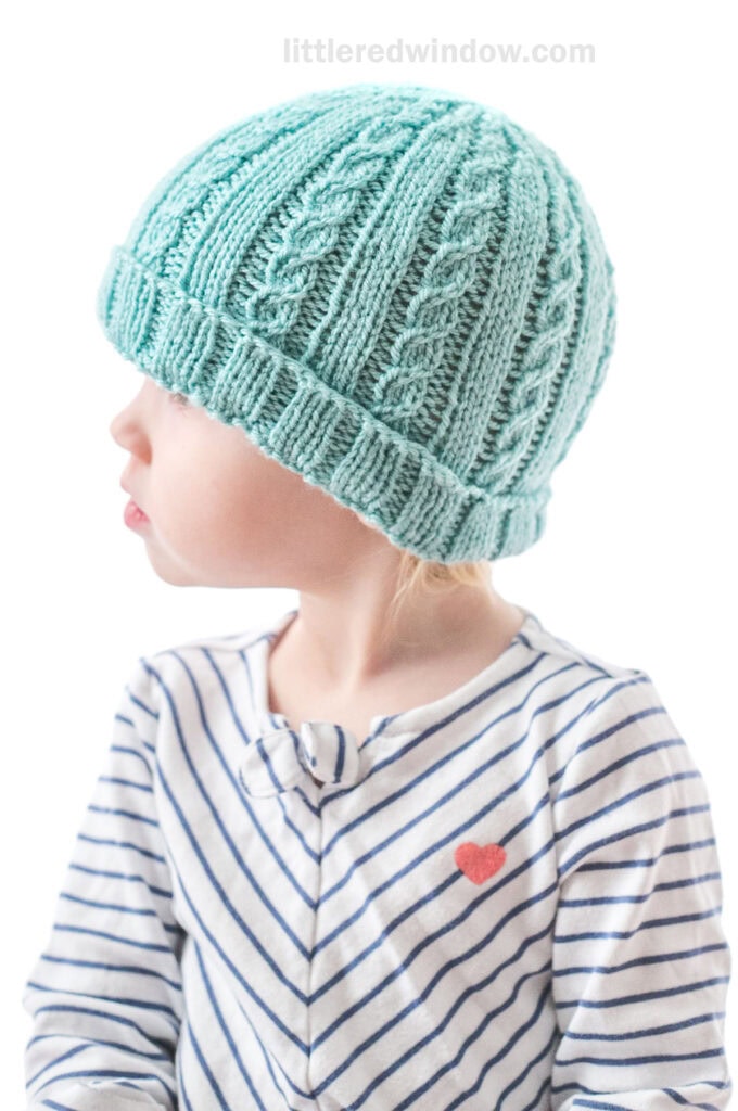 little girl in diagonal striped shirt wearing teal knit hat with alternating plain and cabled ribs looking off to the left