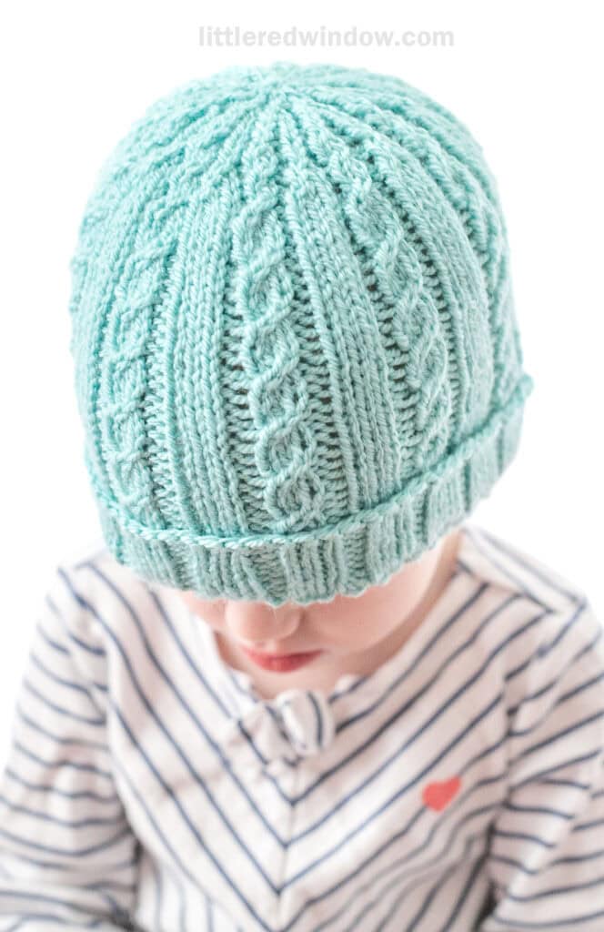 little girl in diagonal striped shirt wearing teal knit hat with alternating plain and cabled ribs looking down