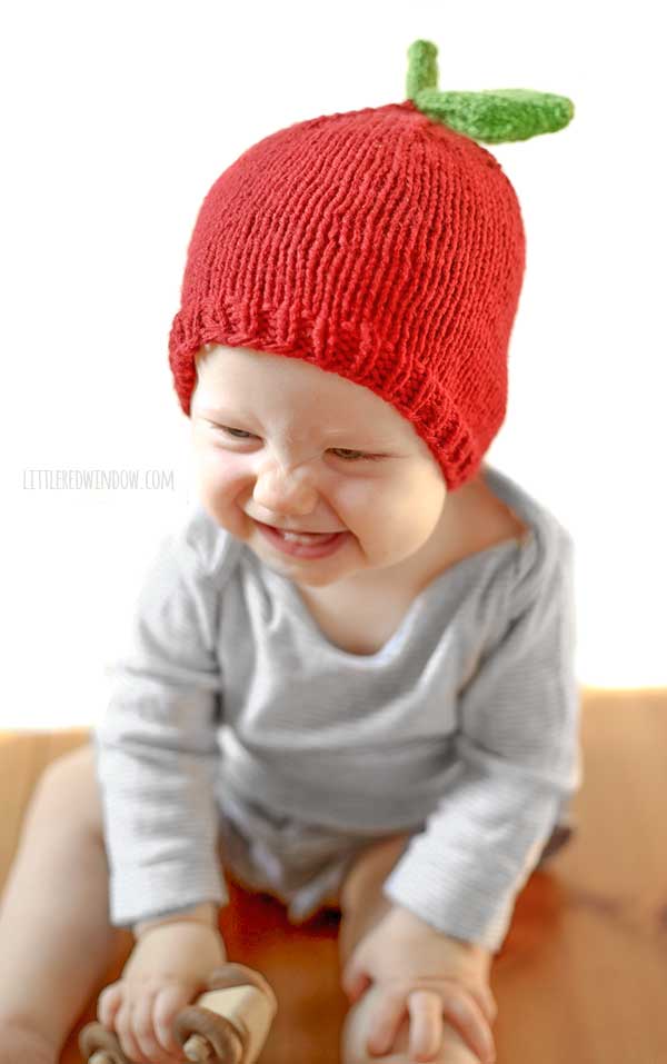 grinning baby in gray shirt wearing a red knit hat that looks like and apple and leaning forward as they sit