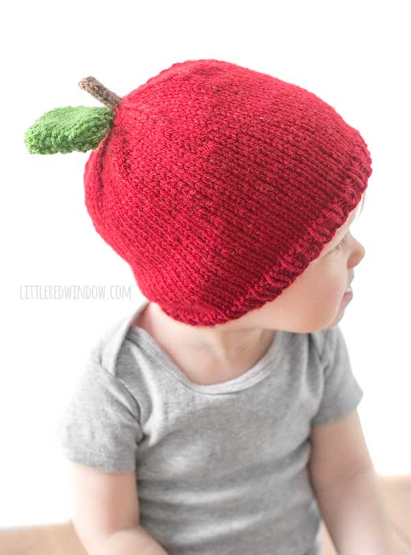 baby in gray shirt wearing a red knit hat that looks like an apple with a brown stem and single green leaf on top sitting on a wood surface and looking off to the right in front of a white background