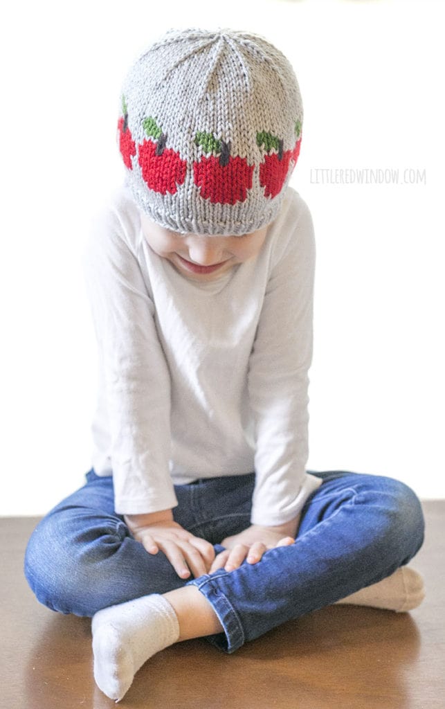 Little girl sitting down and wearing gray knit hat with red apples around the middle