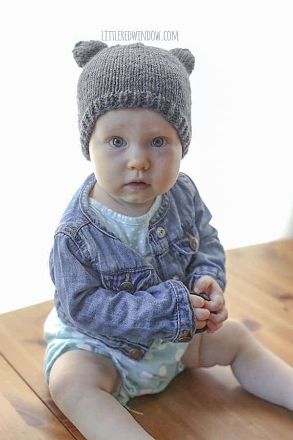 Baby looking at the camera surprised holding a lens cap and wearing a knit hat with bear ears on a wood table in front of a white background