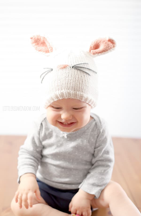 grinning baby in gray shirt wearing a white knit hat with pink and white bunny ears on the top sitting on a wood surface in front of a white background