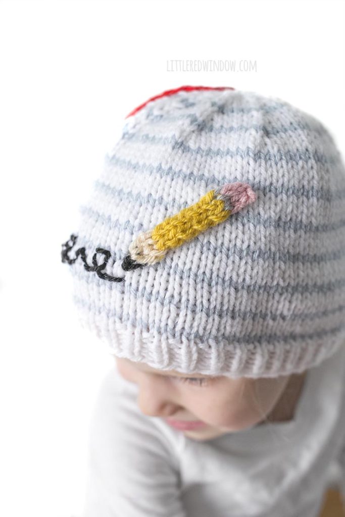 Little girl smiling and wearing a hat that looks like a piece of notebook paper with a pencil scribbling on it and looking down and to the left