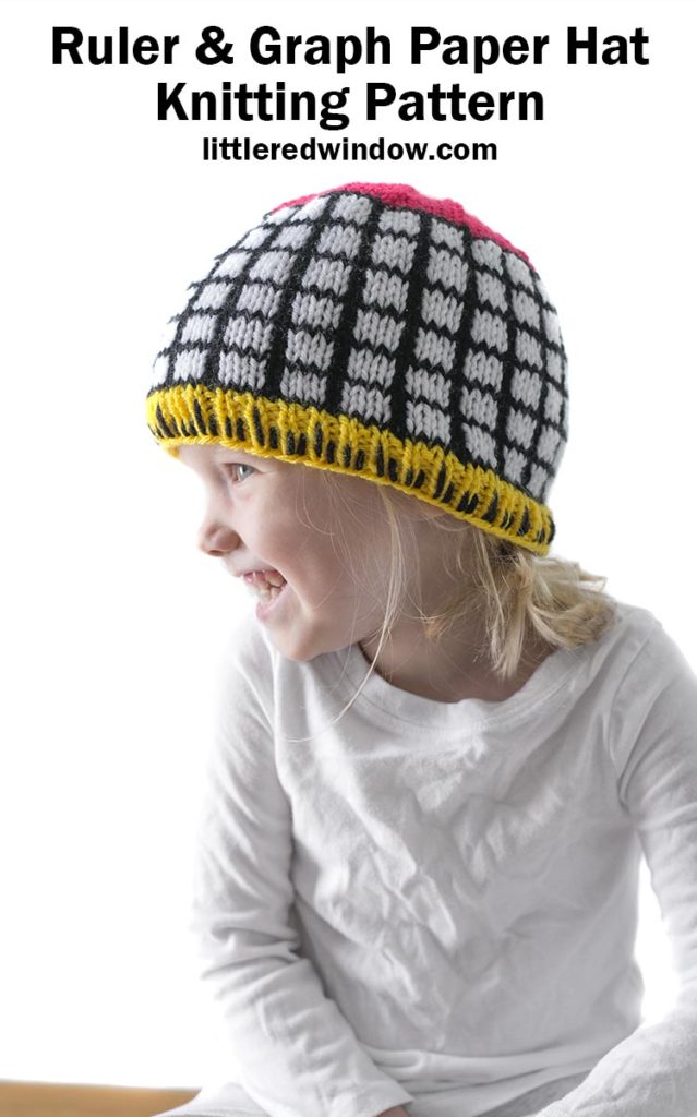Little girl wearing black and white grid knit hat with yellow ruler brim and pink top looking to the left