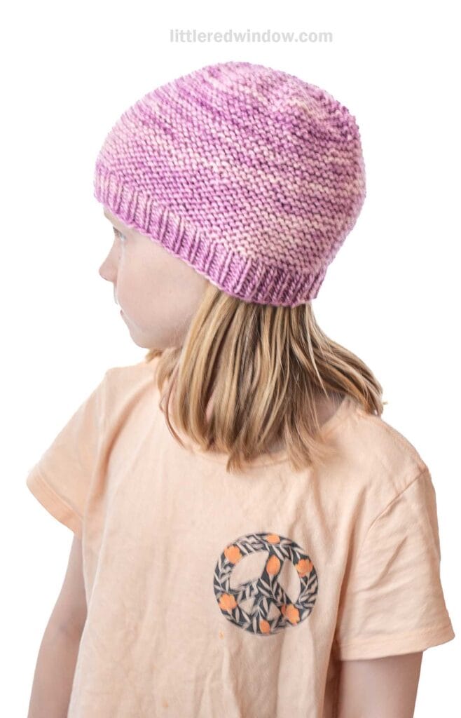A young child with blonde hair wears a pink and white garter stitch hat and a light peach t-shirt with a patterned peace sign design, looking to the left against a white background.