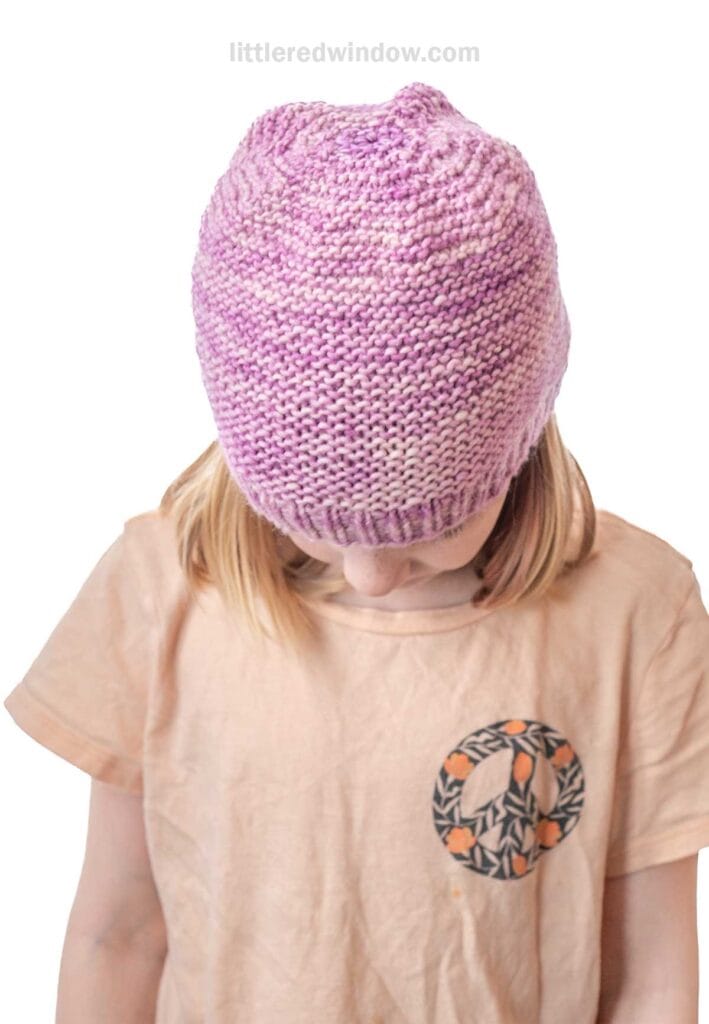 A child wearing a light pink t-shirt with a circular design looks down, showing off a hand-knitted, garter stitch hat in purple and white marbled yarn.