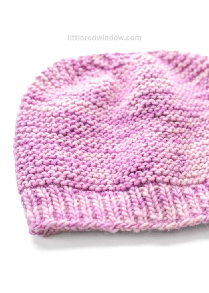 A hand-knitted garter stitch hat for babies in light and dark pink yarn, featuring a ribbed brim and textured stitches, displayed on a white background.