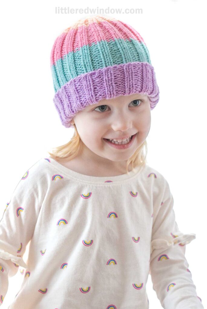 A smiling young child with light hair wears a colorful knit hat made from a vibrant baby hat knitting pattern featuring wide stripes of pink, blue, green, and purple. They are dressed in a cream-colored shirt adorned with small rainbow patterns. The background is plain and well-lit.