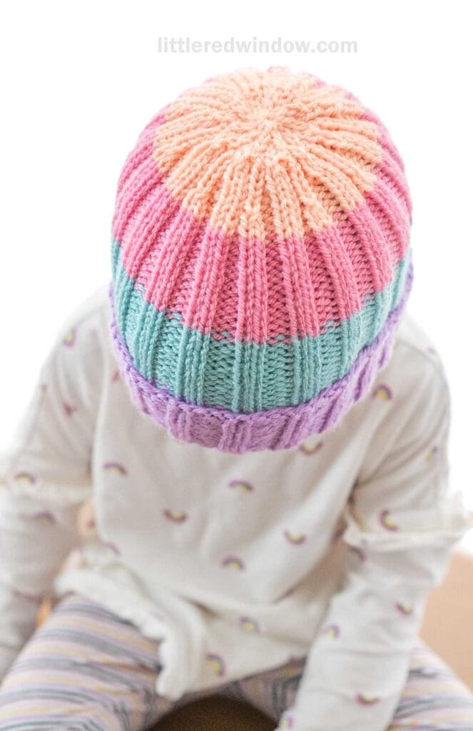 A child wearing a bright, multicolored knit beanie with shades of orange, pink, blue, and purple. They are also dressed in a light-colored, long-sleeved shirt with small rainbow patterns and striped leggings. The text