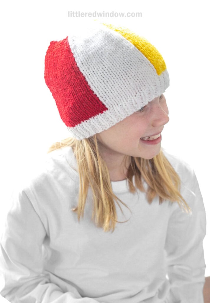 smiling child in white shirt wearing a knit beach ball hat with white red and yellow sections showing looking off to the right