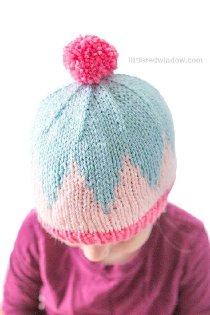 A child wears a big zig hat—a knitted cap with bold blue and pink zigzag patterns, topped with a pink pom-pom—and a purple shirt. The background is white.