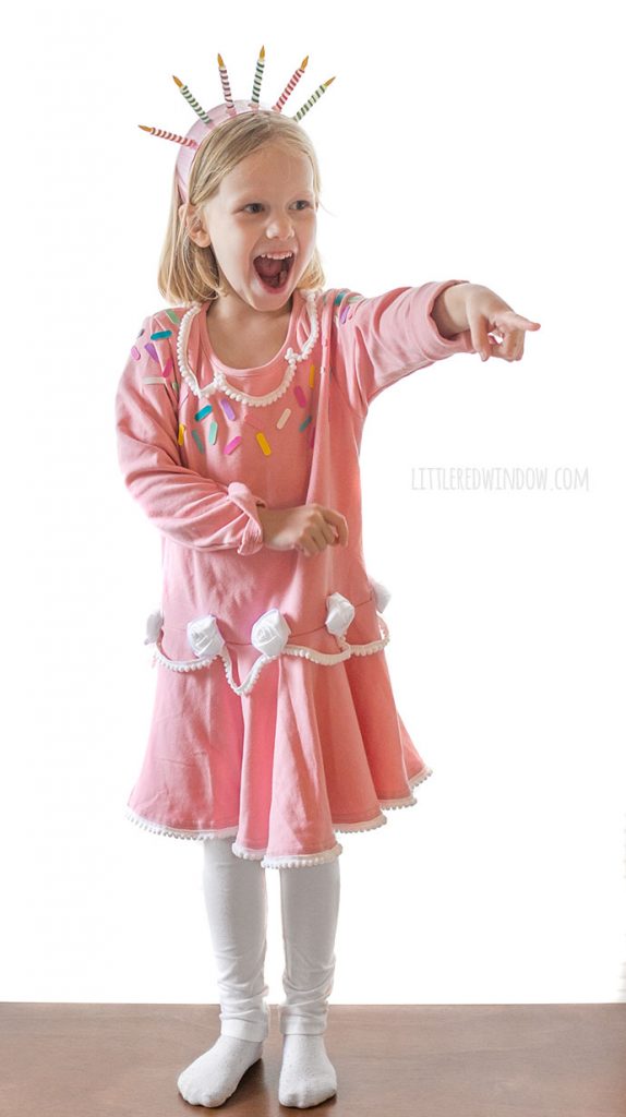 girl in pink birthday cake costume looking surprised and pointing off to the right