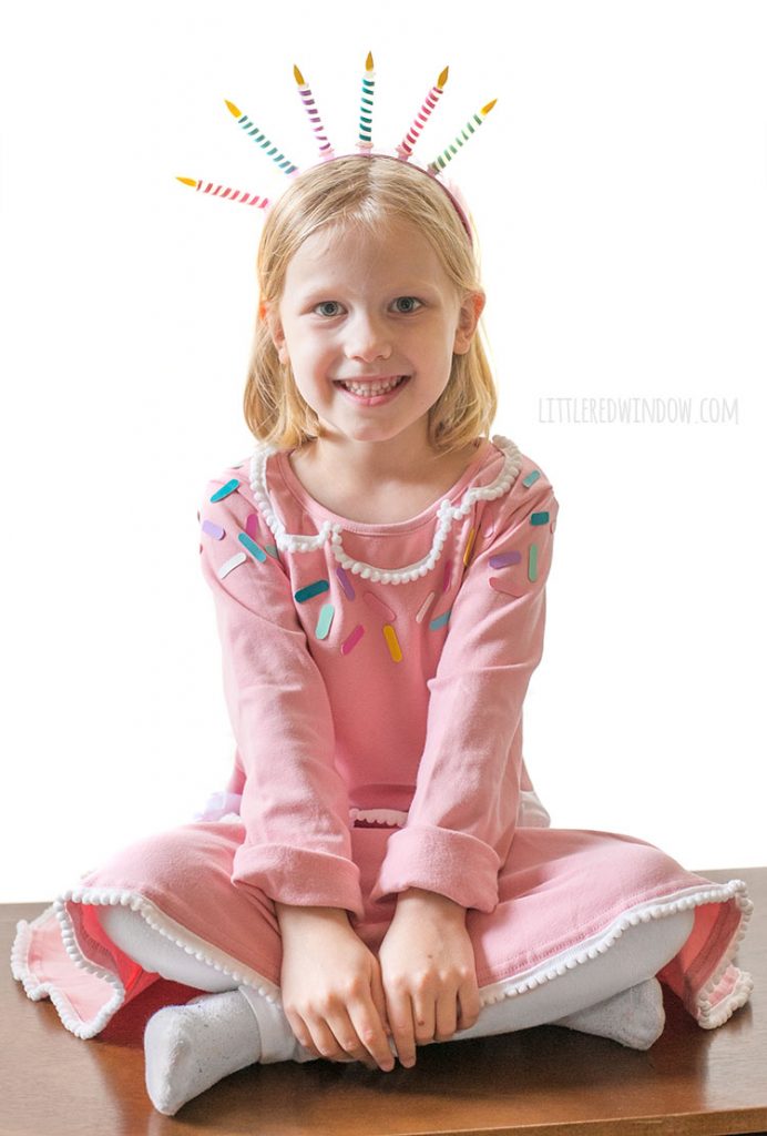 little girl in pink birthday cake costume sitting cross legged and smiling at the camera
