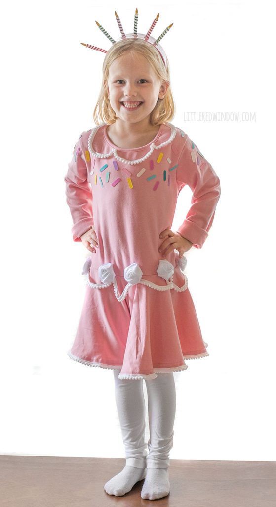smiling girl in pink birthday cake dress with white leggings standing on a table with her hands on her hips.