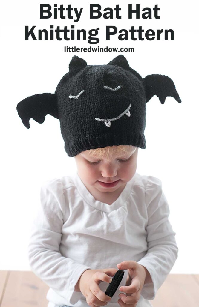 A young toddler wearing a charming black knit bat hat with wings, white eyes, and a smiling mouth with fangs holds an object while looking down. Text above reads 