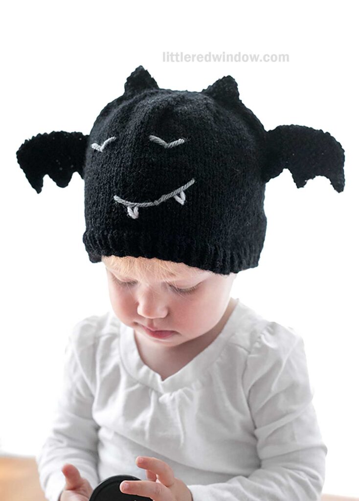 A young child is wearing a black bat hat designed to look like a bat, complete with wings and a face. The child is looking down, wearing a white long-sleeve shirt, and holding an item in their hands. The background is plain white.