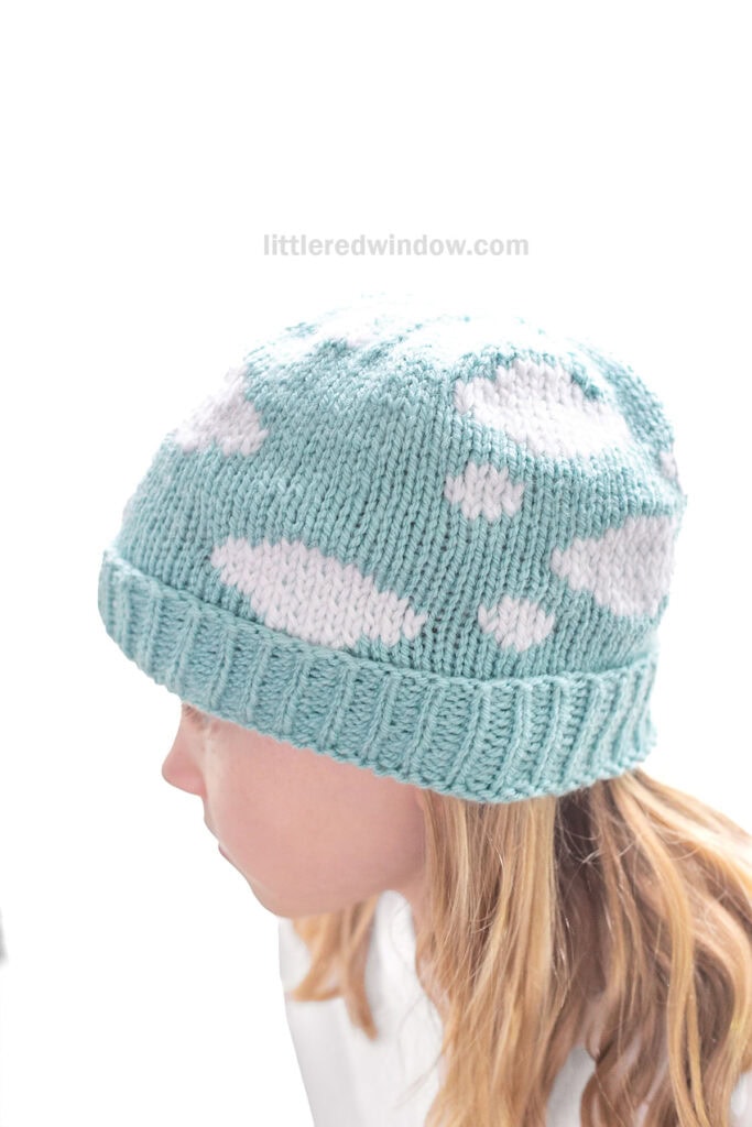 closeup of child wearing light blue knit hat with white clouds all over it in front of a white background