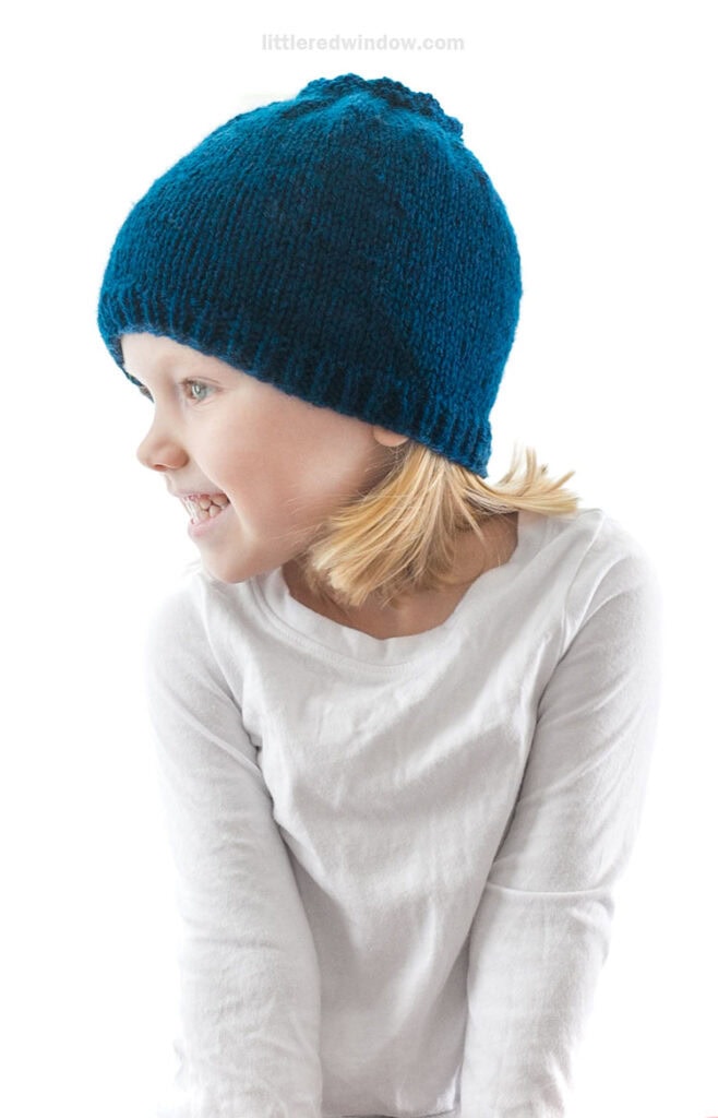 little girl in white shirt looking off to the left and wearing a blue knit hat that looks like a blueberry