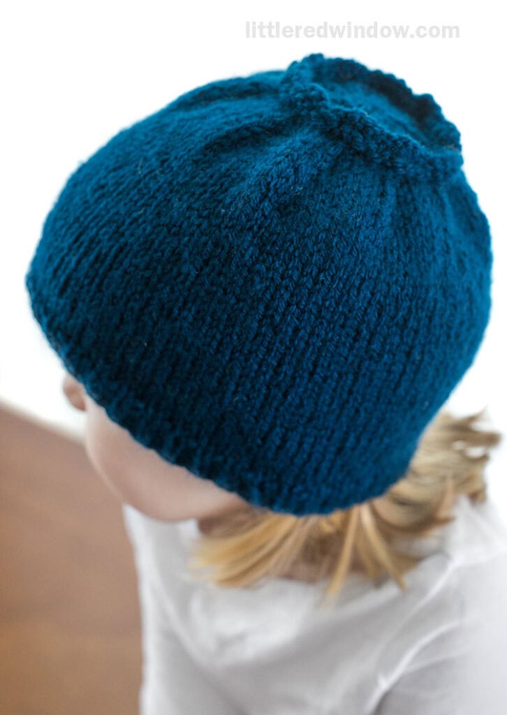 Over the shoulder top view of little girl in white shirt wearing a blue knit hat that looks like a blueberry