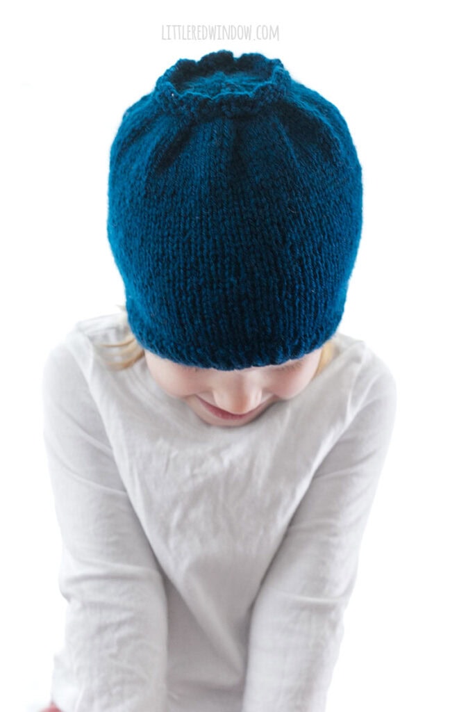 little girl in white shirt looking down and wearing a blue knit hat that looks like a blueberry