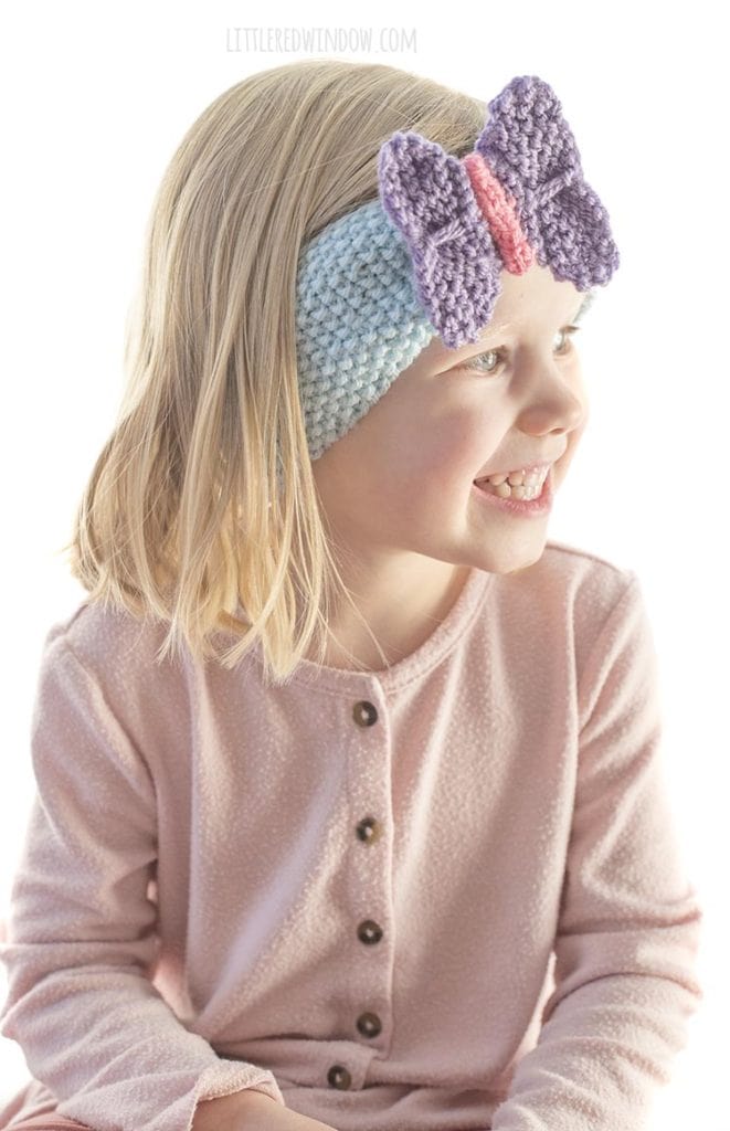 little girl in pink shirt wearing blue knit headband with purple butterfly on the front looking off to the right