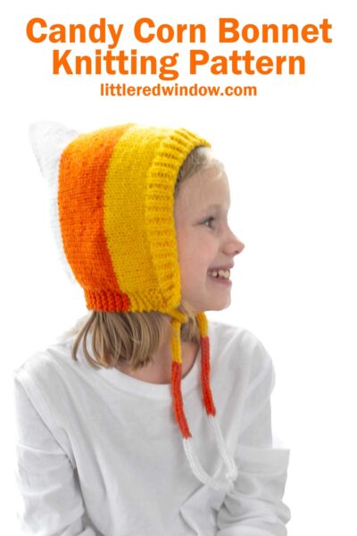A child wearing a knitted bonnet resembling candy corn in colors of white, orange, and yellow. They are smiling and looking to the side. Text above reads, "Candy Corn Bonnet Knitting Pattern" followed by "littleredwindow.com.