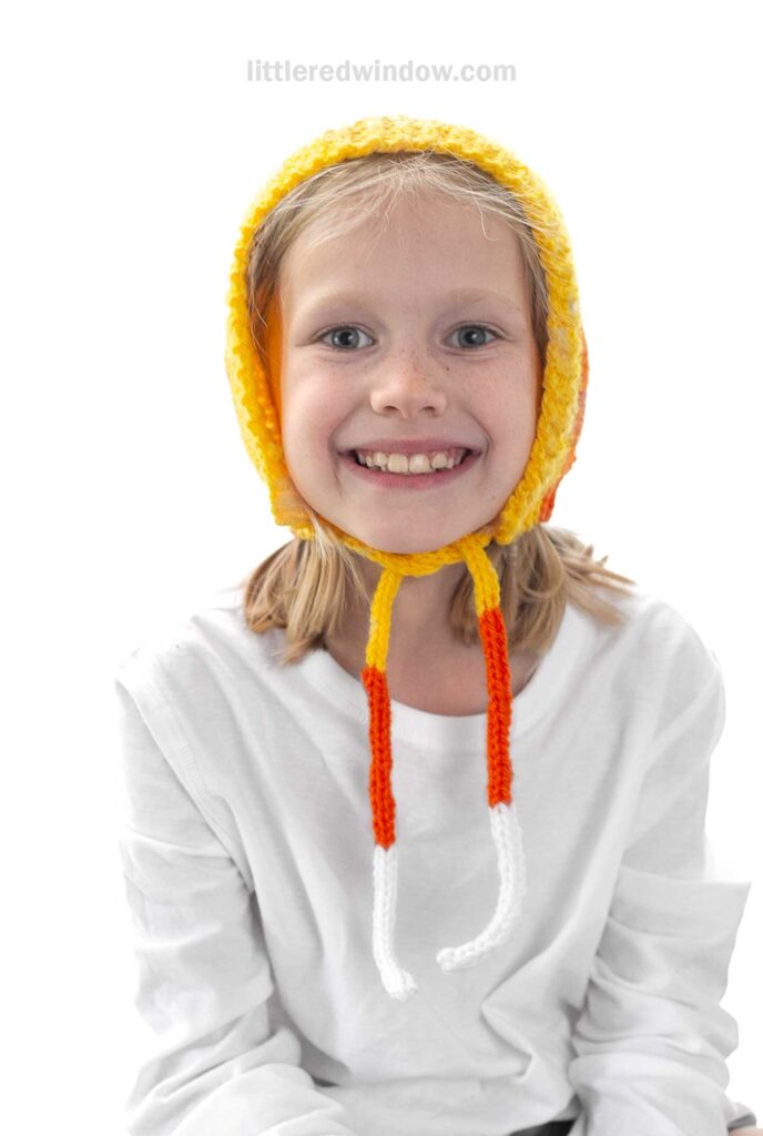 A smiling child wearing a handmade knit hat with yellow, orange, and white stripes. The hat covers their head and part of their forehead, with two braided tassels hanging down. The child is wearing a white long-sleeve shirt and sitting against a white background.