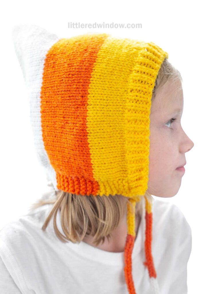 A child with blonde hair wears a handmade knit hat featuring vertical stripes in orange, yellow, and white colors. The child's face is in profile, looking to the right, against a plain white background. The hat has two dangling ties.