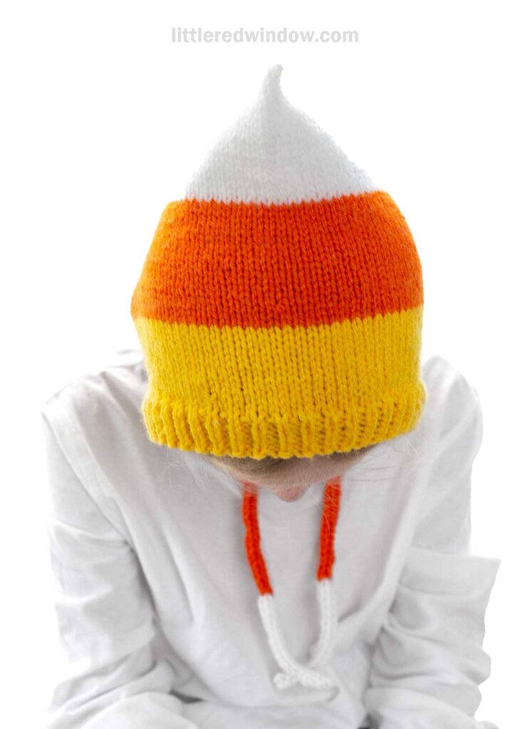 A person wearing a knitted hat designed to look like candy corn, featuring white, orange, and yellow sections. The person is dressed in a white shirt with orange and white braided strings and is looking down. The background is white.
