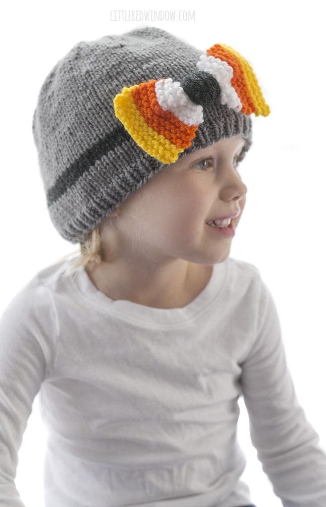 little girl in white shirt looking off to the right and wearing a gray knit hat with candy corn colored bow on the front