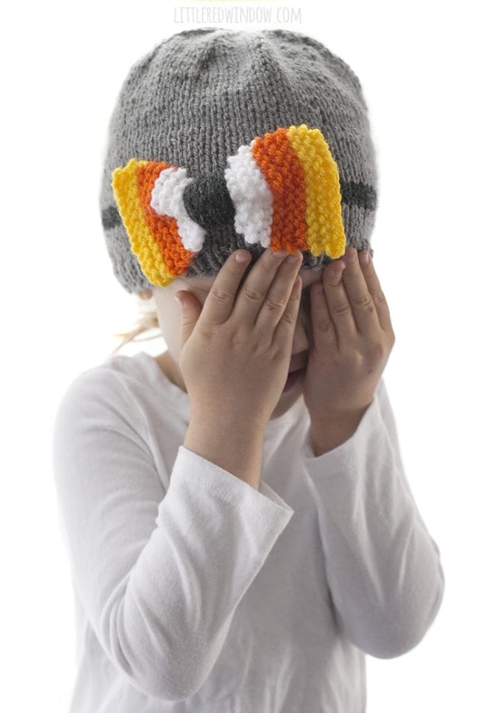 little girl with hands over face wearing a gray knit hat with candy corn colored bow on the front