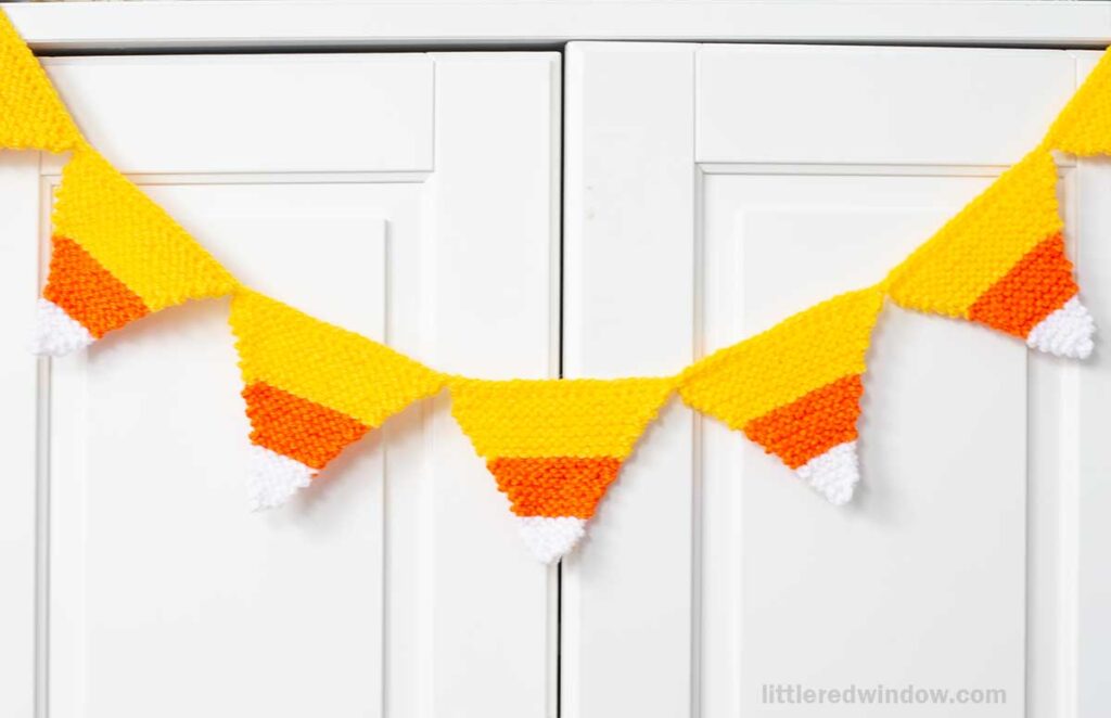 Candy corn garland, knitted in white, orange, and yellow stripes, is hanging across white cabinet doors.