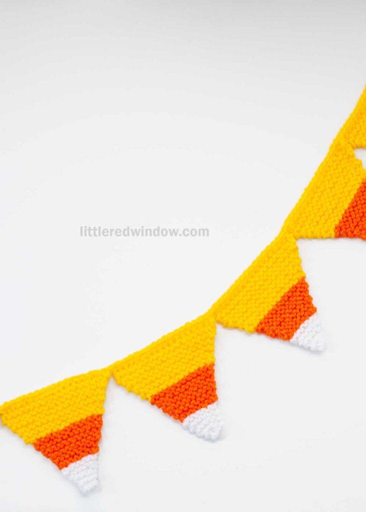 A candy corn garland of knitted triangular pennants in yellow, orange, and white hangs against a plain white background.
