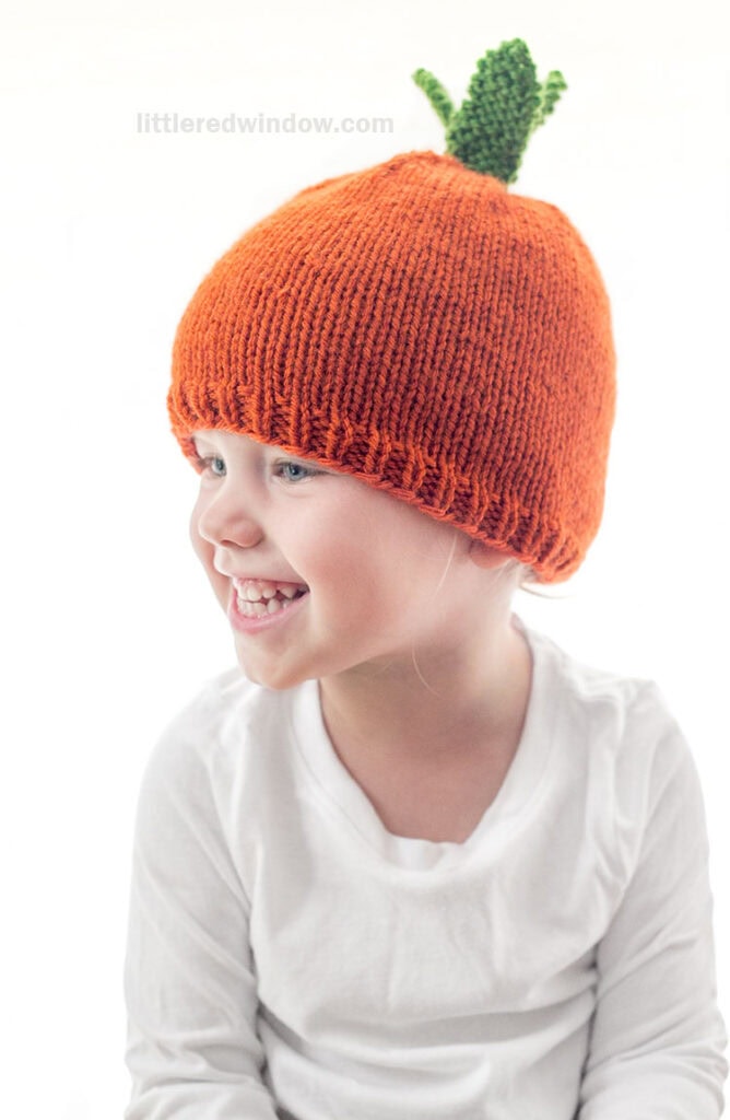 child in white shirt wearing an orange knit carrot hat with green knit carrot leaves on top looking slightly to the left and laughing