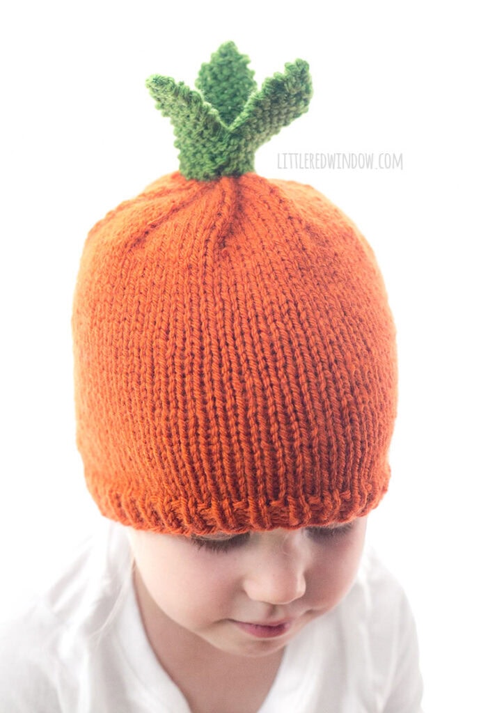 child in white shirt wearing an orange knit carrot hat with green knit carrot leaves on top looking down to the right