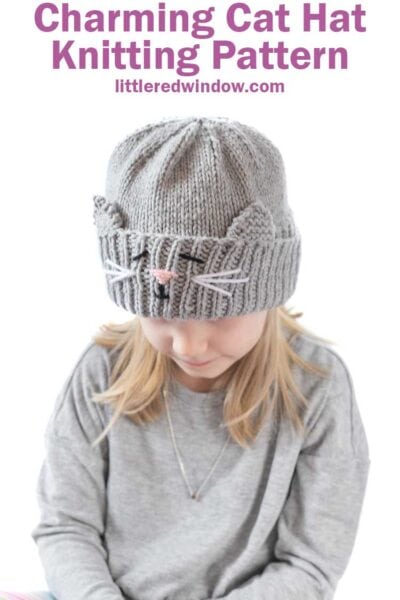 blond girl in gray shirt looking down at her lap and wearing a gray knit hat with a cat face and ears embroidered into the thick ribbed folded brim