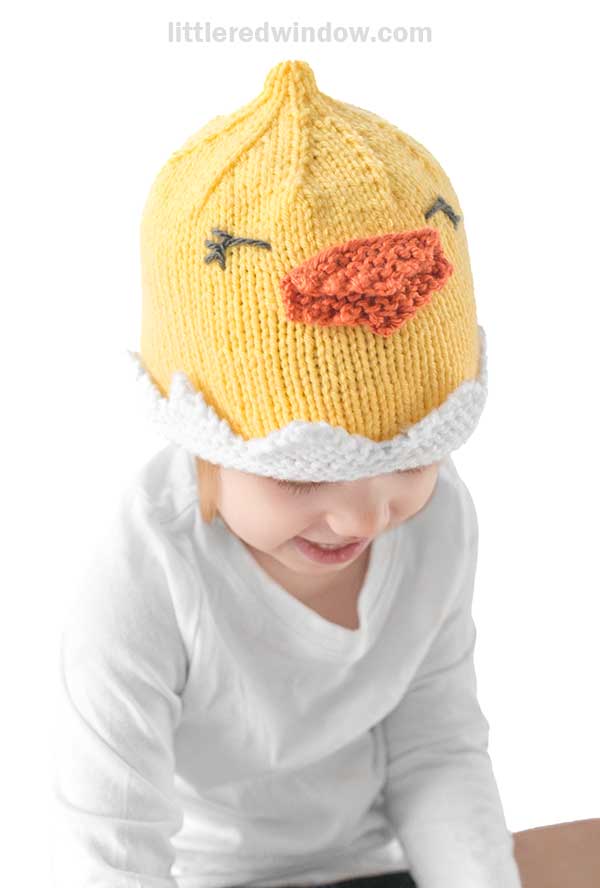 A child is beaming in a charming chick hat—a knitted yellow masterpiece with an orange beak and black embroidered eyes. Paired with a white long-sleeve shirt, they stand against a simple white backdrop, radiating joyful innocence.