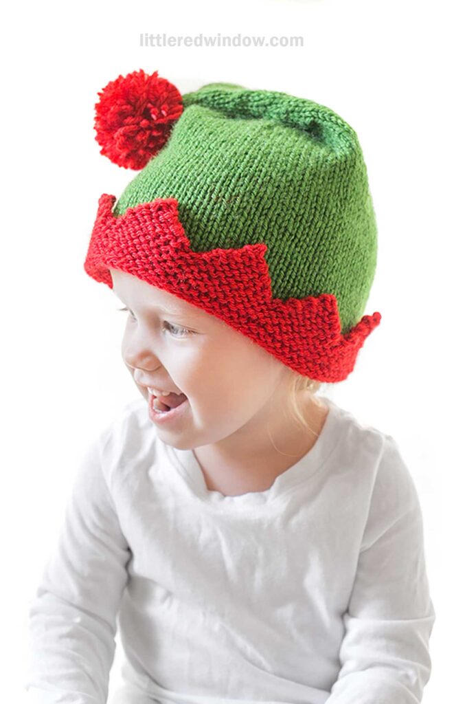 The child, donning an elf hat with a delightful red zigzag edge and a playful pom-pom on top, is grinning widely in their white shirt. This charming knitted accessory adds a whimsical touch to their cheerful look.