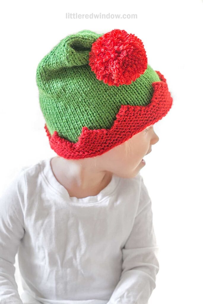 A child wearing an elf hat with a green and red zigzag knitting pattern sits, gazing off to the side. The playful pom-pom atop the hat adds charm to their look, complemented by a simple white shirt. The minimalist white background keeps focus on the child's festive attire.