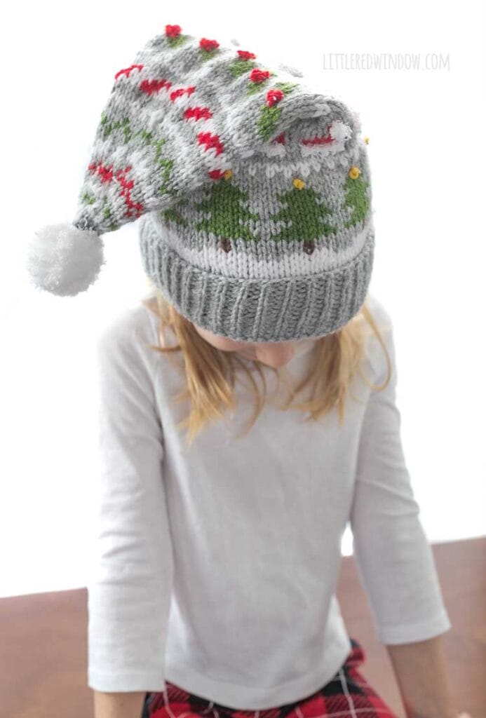 blond girl in white long sleeved shirt wearing a gray knit stocking cap decorated with patterns of Christmas trees Santa hats holly berries and candy cane stripe and a white pom pom on top looking down with stocking cap to the front